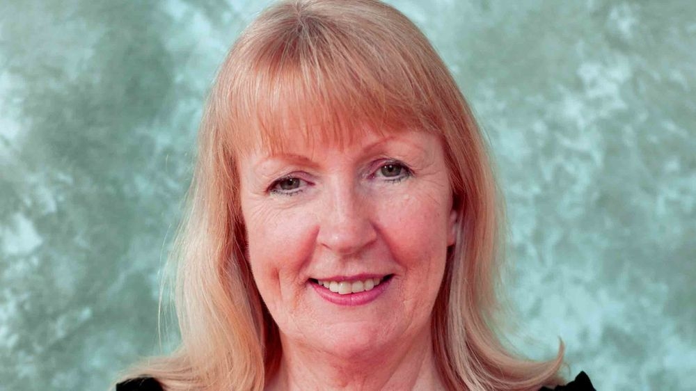 Adele Gibson, head and shoulders, smiling at the camera against a blue-green studio backdrop