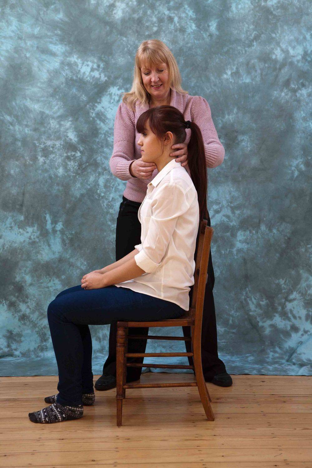 Adele standing behind a seated pupil on a plain wooden chair, both hands lightly at the pupil's neck and head