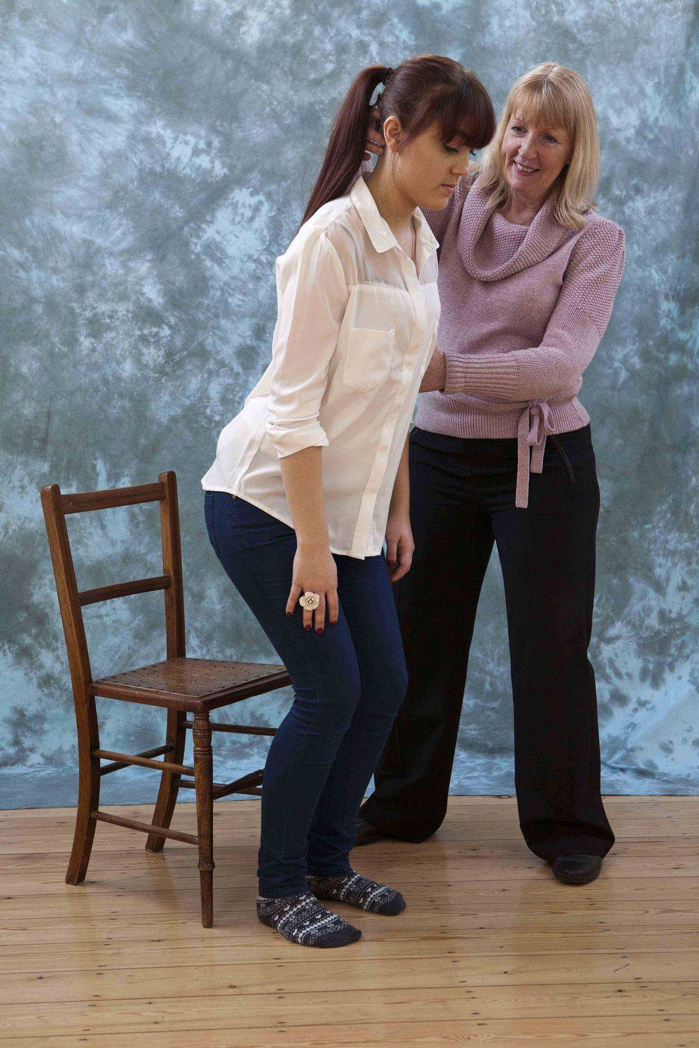 The pupil half-standing beside the empty wooden chair, Adele alongside guiding her